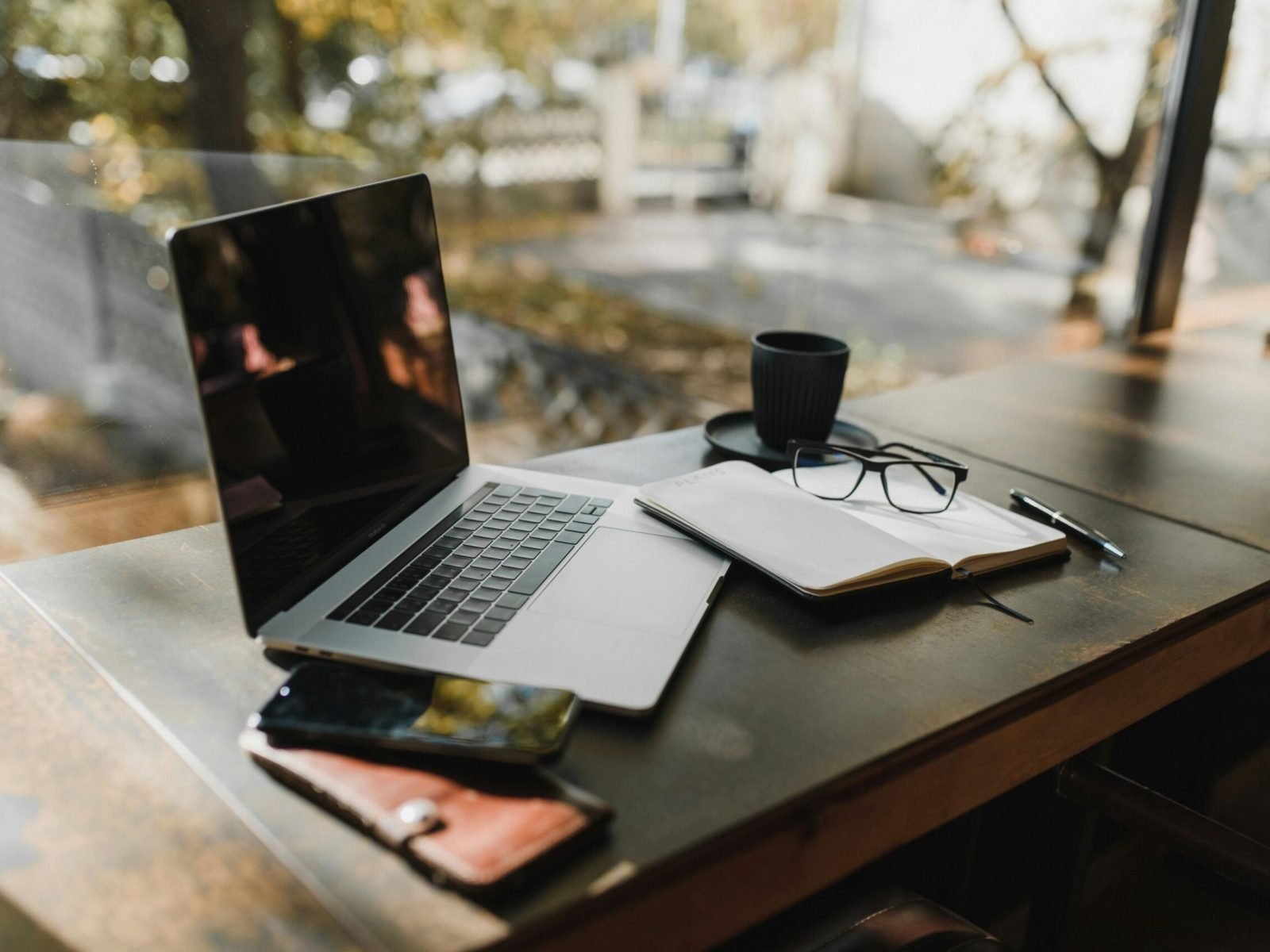 A serene office setup with a laptop, notebook, and coffee near a window, reflecting an autumn scene.