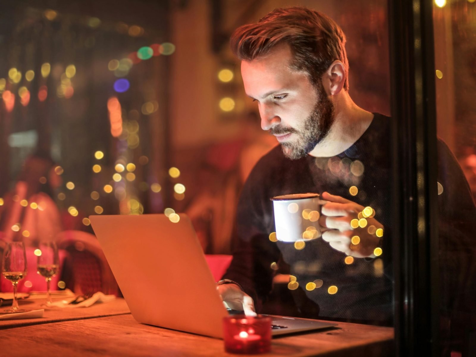 A young man with a beard is working on a laptop at a cozy cafe with warm lighting, holding a coffee cup.